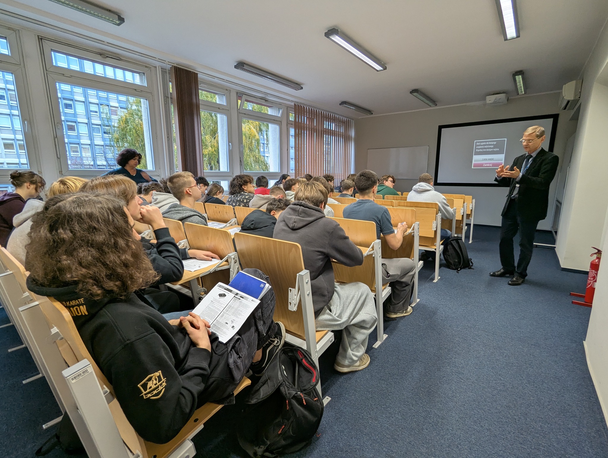 Zdjęcie przedstawia mgr Aleksandra Kubota, wykładowcę Centrum Języków i Komunikacji podczas zajęć ze studentami i uczniami w sali wykładowej.